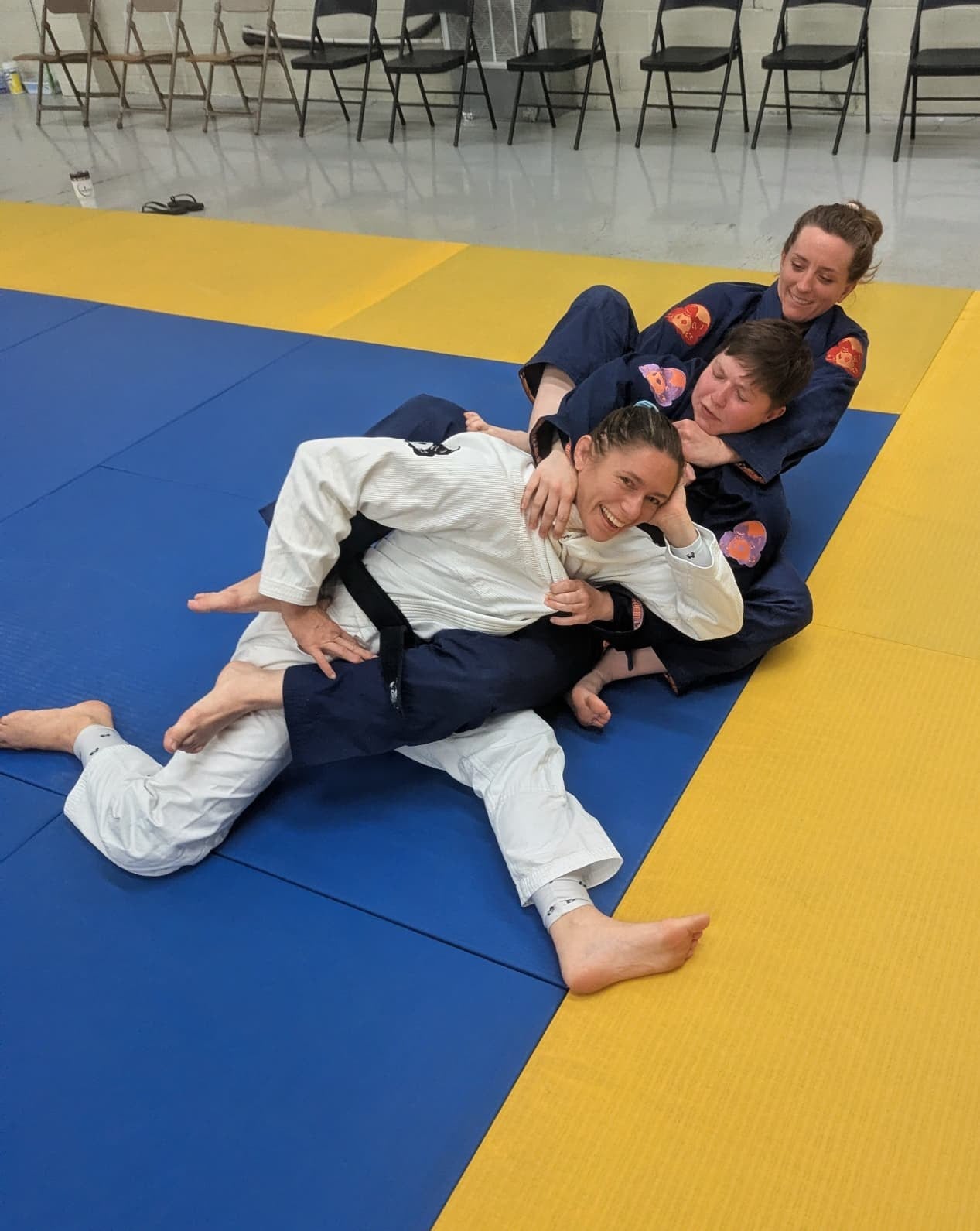 Three training partners piled up laughing on the mats, two in navy Panda gis